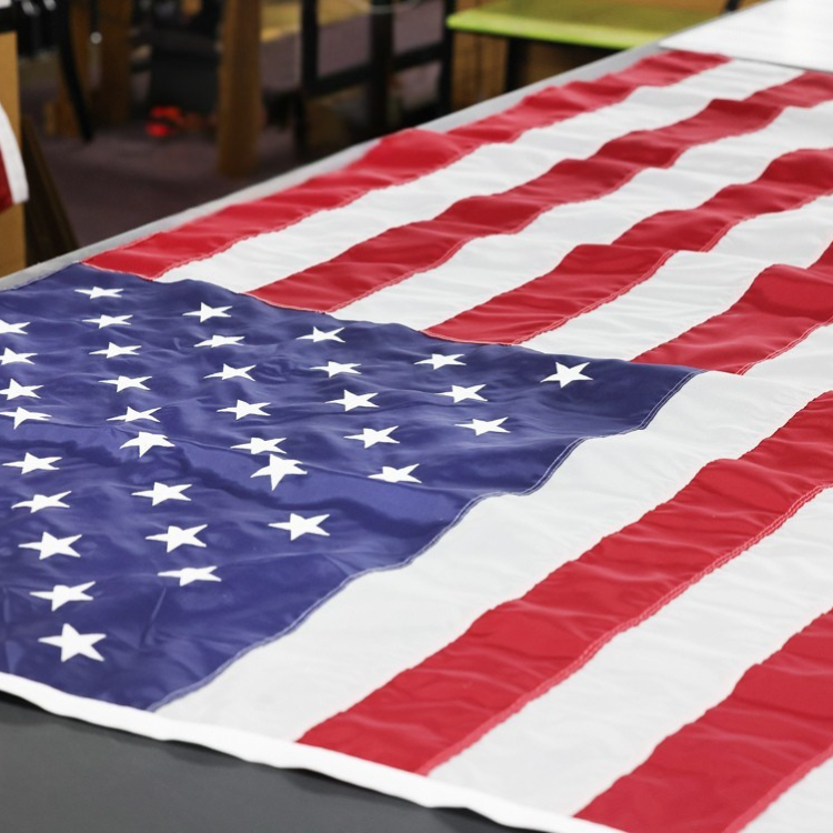 Printed American flag waving against a blue sky, featuring red and white stripes and a blue field