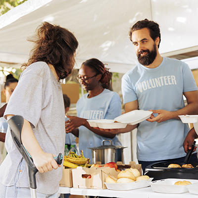 Photograph showing a serving line, with a man handing a plate of food to a woman who has a forearm cruth.